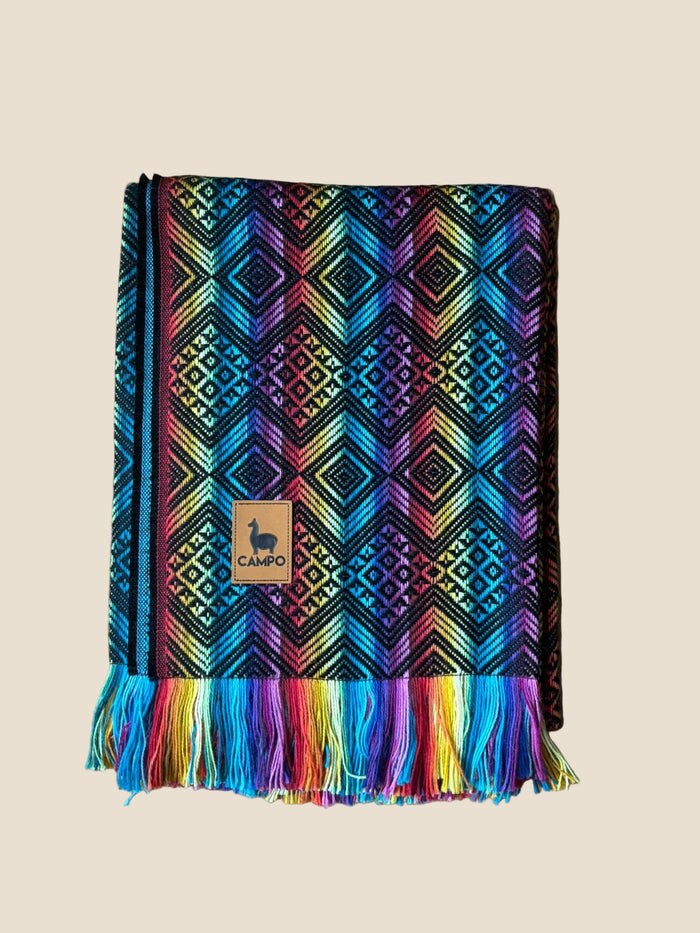Colorful patterned blanket with fringes on a wooden surface, featuring a 'CAMPQ' brand label.