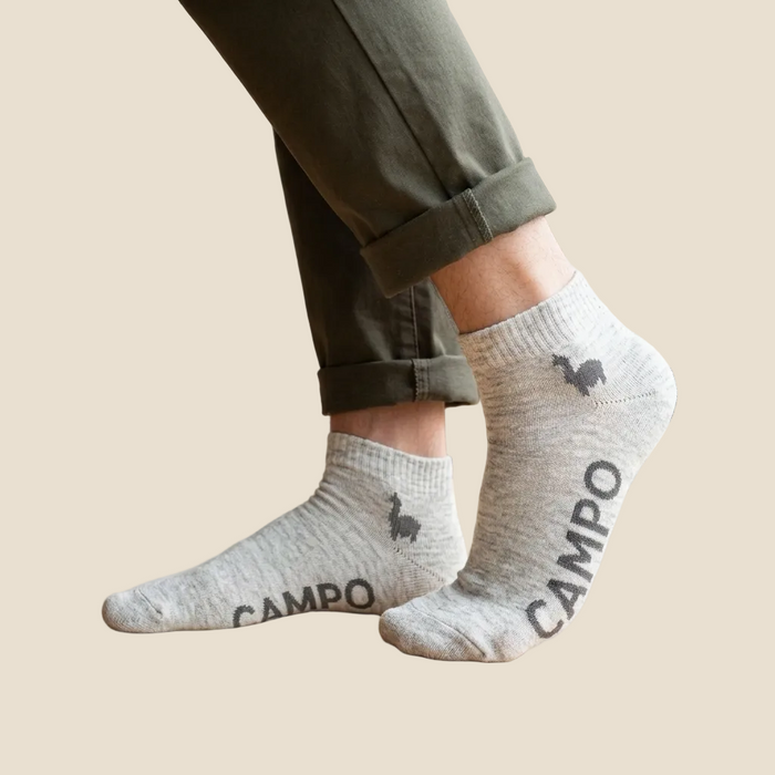 A person wearing light heather gray ankle socks with dark gray 'CAMPO' text and a small rooster silhouette, paired with olive green pants, standing on a light wood floor near a window.