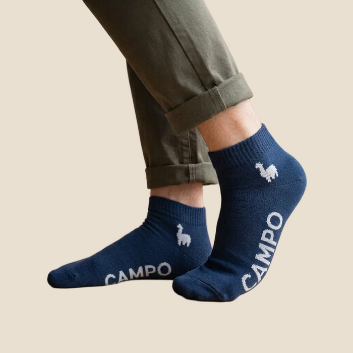 A person wearing navy blue ankle socks with white llama graphics and the brand name 'CAMPO' printed on the side, paired with olive green pants in a bright indoor setting with a wooden floor and window.