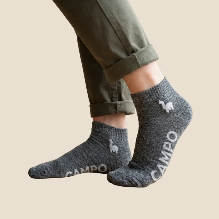 A person wearing dark heather gray ankle socks with white branding and llama graphics, paired with olive green pants, standing on a light wood floor near a window.