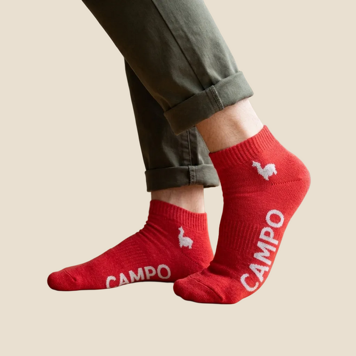 Red low-cut ankle socks with white 'CAMPO' text and llama graphics, worn with cuffed olive green pants against a light beige background.