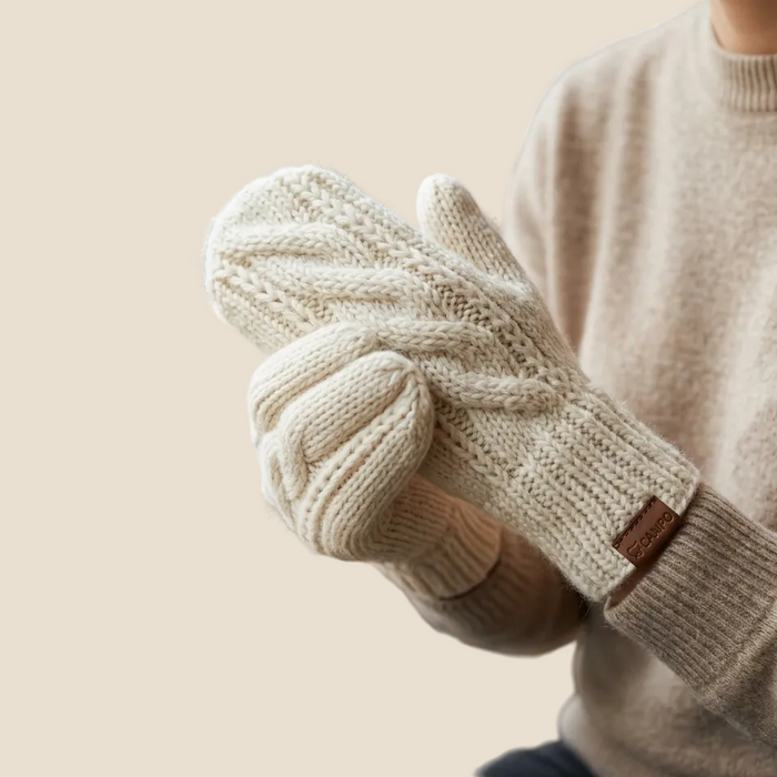 Person wearing beige knitted gloves with a wooden tag, sitting at a wooden table with a mug.