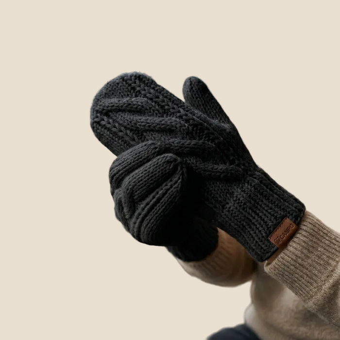 Person wearing dark gray knit gloves with a beige sweater, sitting by a window with a mug on a wooden table.