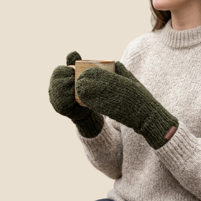 Person wearing green knitted gloves holding a wooden mug against a beige background