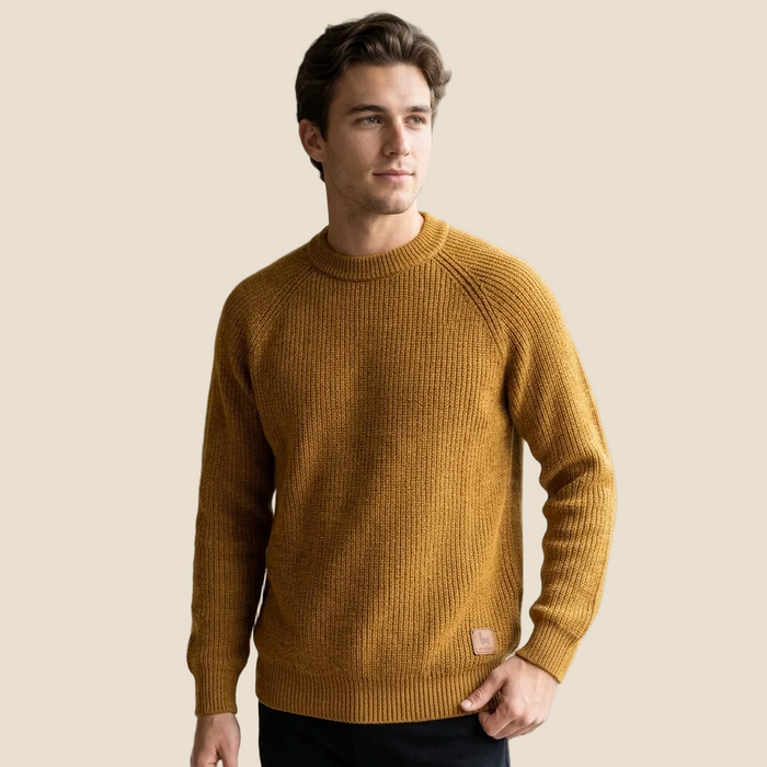 A man wearing a ribbed knit crewneck sweater in a warm mustard yellow color, paired with dark pants, set against a solid light beige background.