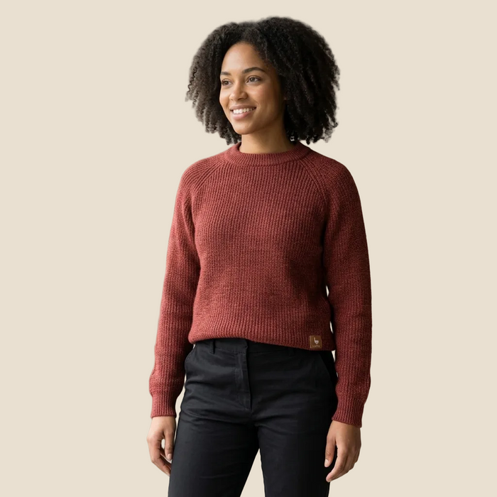 A woman with curly black hair wearing a ribbed knit crewneck sweater in a dusty rose color and black pants, standing against a solid beige background.