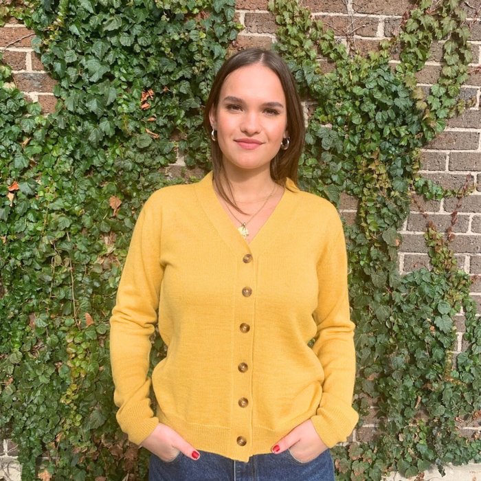 Woman wearing a yellow cardigan standing in front of a brick wall with green ivy