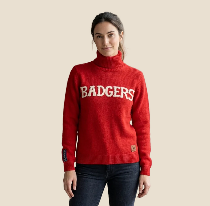 A woman wearing a bright red turtleneck sweater with 'BADGERS' in white varsity lettering across the chest, paired with dark jeans, standing in a modern room with indoor plants.