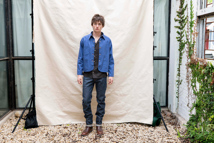 Person wearing a blue jacket and dark jeans standing in front of a white backdrop outdoors.
