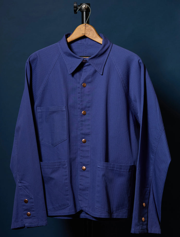 Barista Chore Coat in Washed Herringbone with brown pearl snaps on a hanger against a dark background