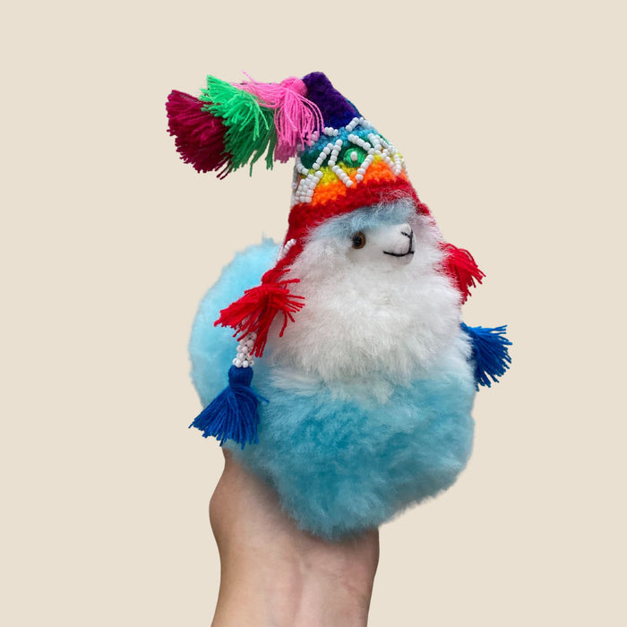 Plush toy llama with colorful hat held by a hand against a beige background