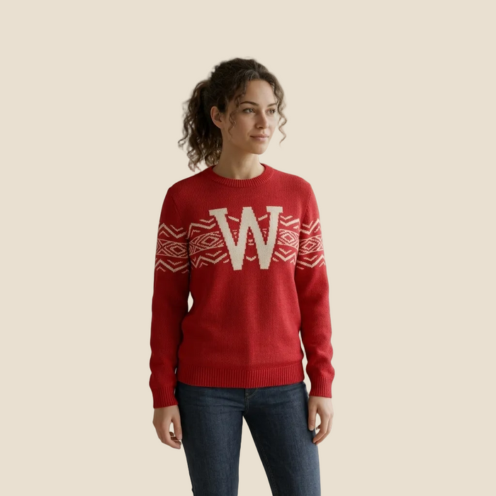 A woman wearing a red crewneck sweater with a large cream letter 'W' and geometric patterns across the chest, paired with dark blue jeans, standing in a modern room with potted plants.