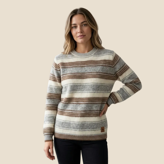 A woman modeling a long-sleeve crewneck sweater with horizontal stripes in beige, brown, and gray, paired with dark pants, against a solid light beige background.