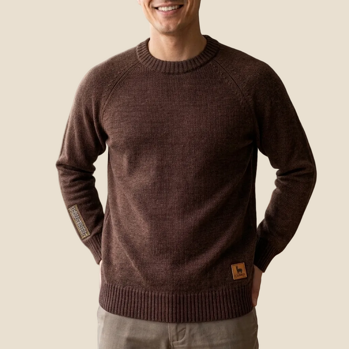 A man wearing a dark brown ribbed knit crewneck sweater with a small brown leather logo patch on the lower hem and a textured sleeve patch, set against a solid beige background.