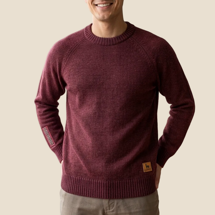 A man wearing a maroon ribbed knit crewneck sweater with a small brown logo patch on the lower hem and a patterned sleeve tag, photographed against a solid light beige background.