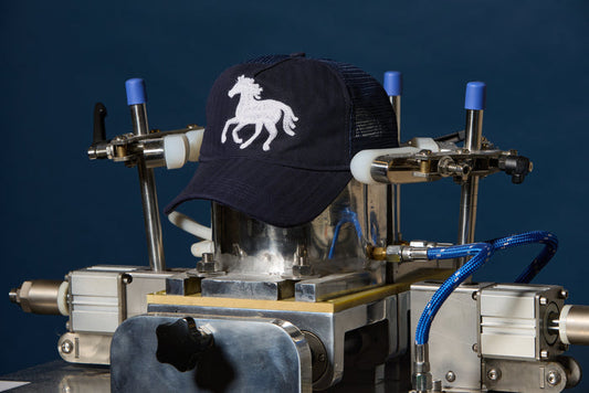 A navy blue trucker cap with a white embroidered horse logo is being held by a metal and pneumatic pressing machine against a dark blue background.