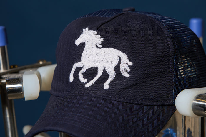 A navy blue trucker cap with a white embroidered horse logo, shown resting on a metal display stand against a dark blue background.