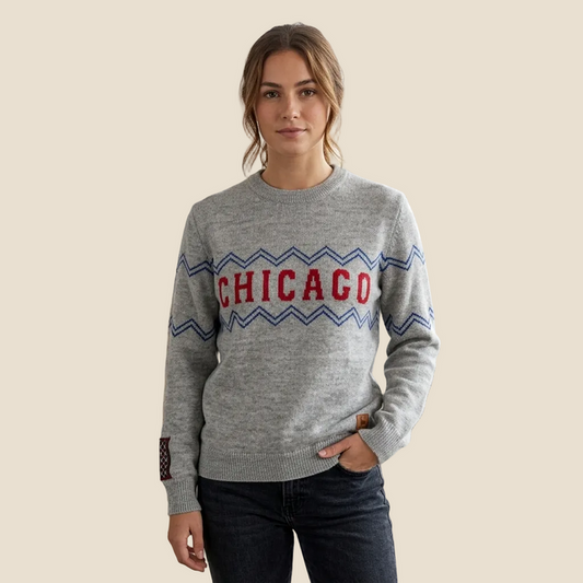A woman wearing a heather gray knit sweater with a red and blue chevron pattern and the word 'CHICAGO' in red across the chest, paired with dark jeans, against a solid beige background.