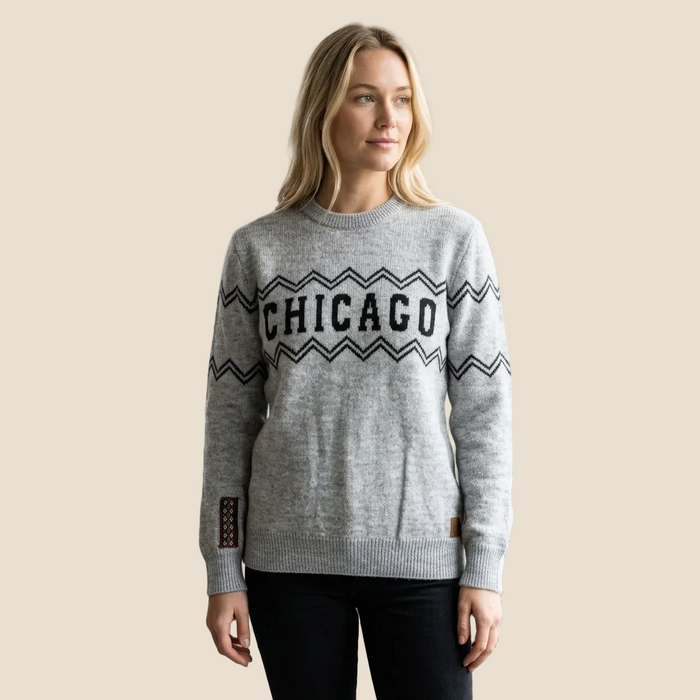 A woman modeling a light gray knit crewneck sweater with 'CHICAGO' in black lettering across the chest, framed by black zigzag patterns, against a solid beige background.
