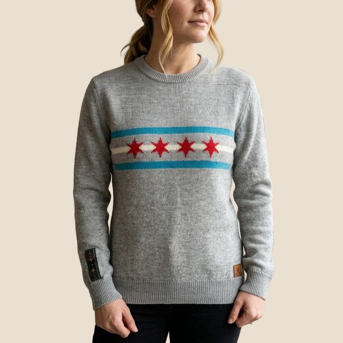 A woman wearing a heather gray crewneck sweater with a Chicago flag-inspired chest pattern featuring red stars and blue and white stripes, set against a solid beige background.