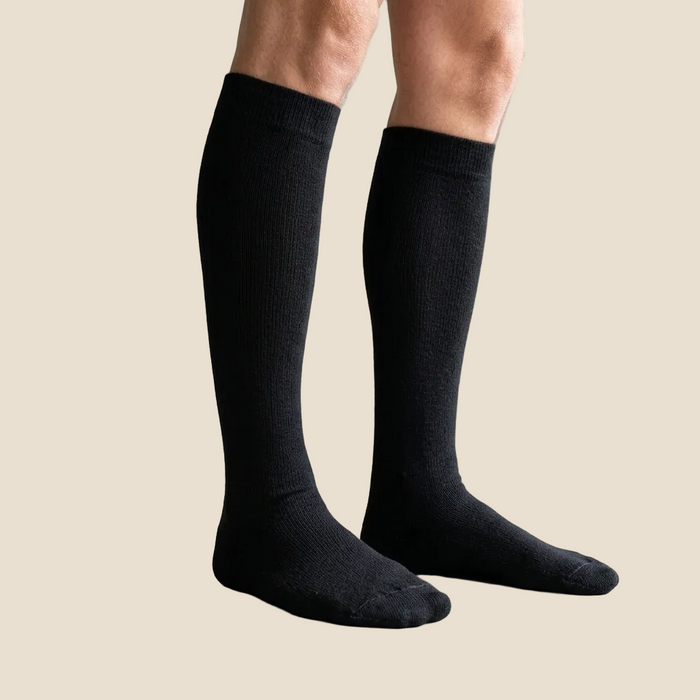 Person wearing black knee-high socks in a minimal indoor setting with a plant and wooden furniture.
