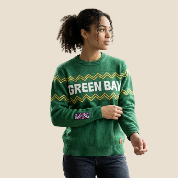 A woman wearing a green crewneck sweater with 'GREEN BAY' in white letters across the chest, decorated with yellow and white chevron patterns and a purple-and-white patch on the sleeve, posed against a solid beige background.