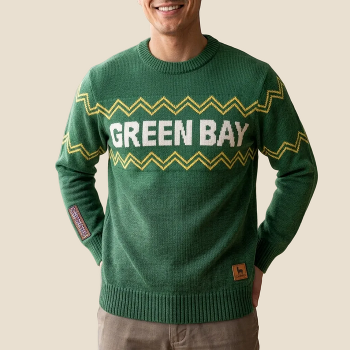 A man wearing a green crewneck sweater with 'GREEN BAY' in white letters across the chest, bordered by yellow zigzag stripes. The sweater also features a small patch on the sleeve and a small brown logo patch near the bottom hem, set against a solid beige background.