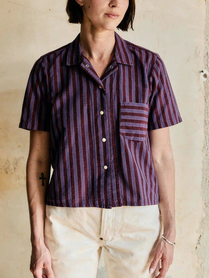 Female wearing a short-sleeve, button-up shirt with purple and red vertical stripes against a neutral background.