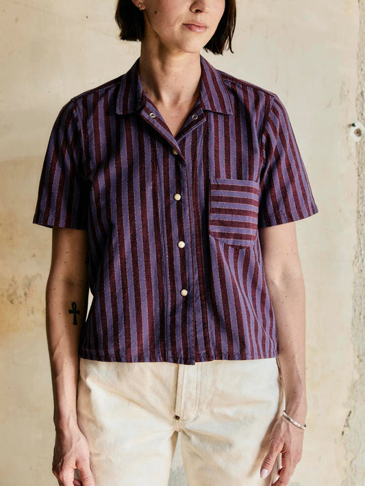 Female wearing a short-sleeve, button-up shirt with purple and red vertical stripes against a neutral background.