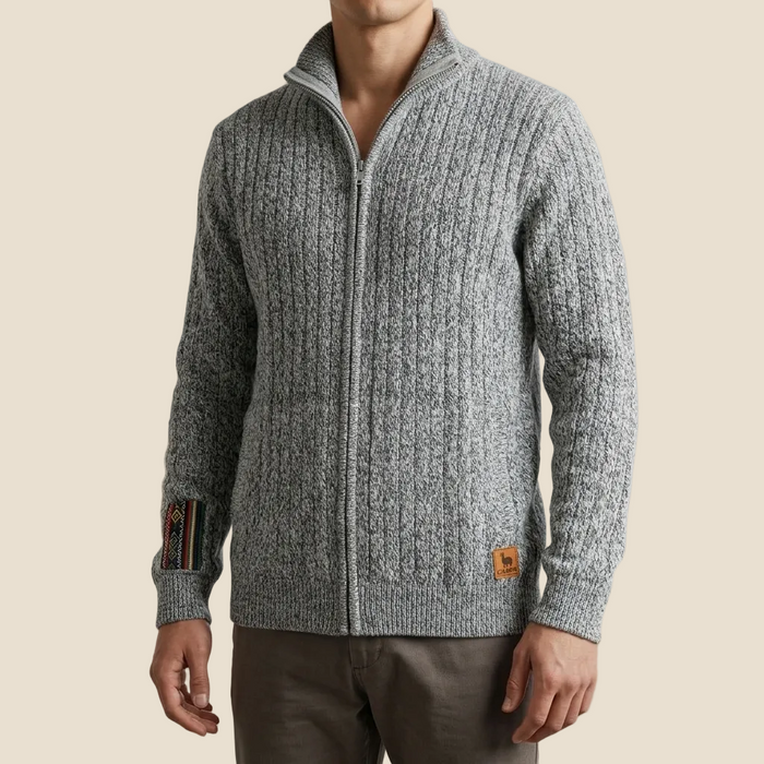 A man wearing a gray cable-knit zip-up sweater with a stand collar, paired with brown trousers, against a solid beige background.