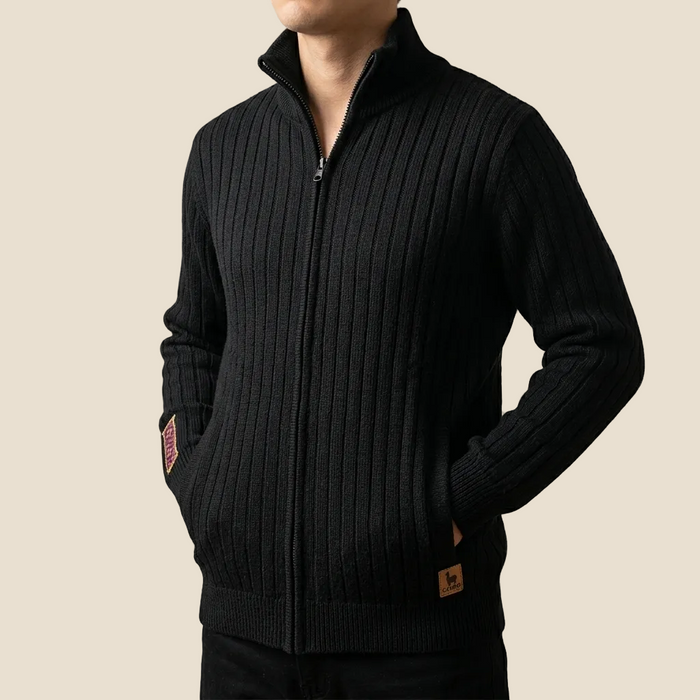 A man wearing a black ribbed knit zip-up sweater with a stand collar, side pockets, and small brown brand patches on the sleeve and hem, shown against a plain beige background.