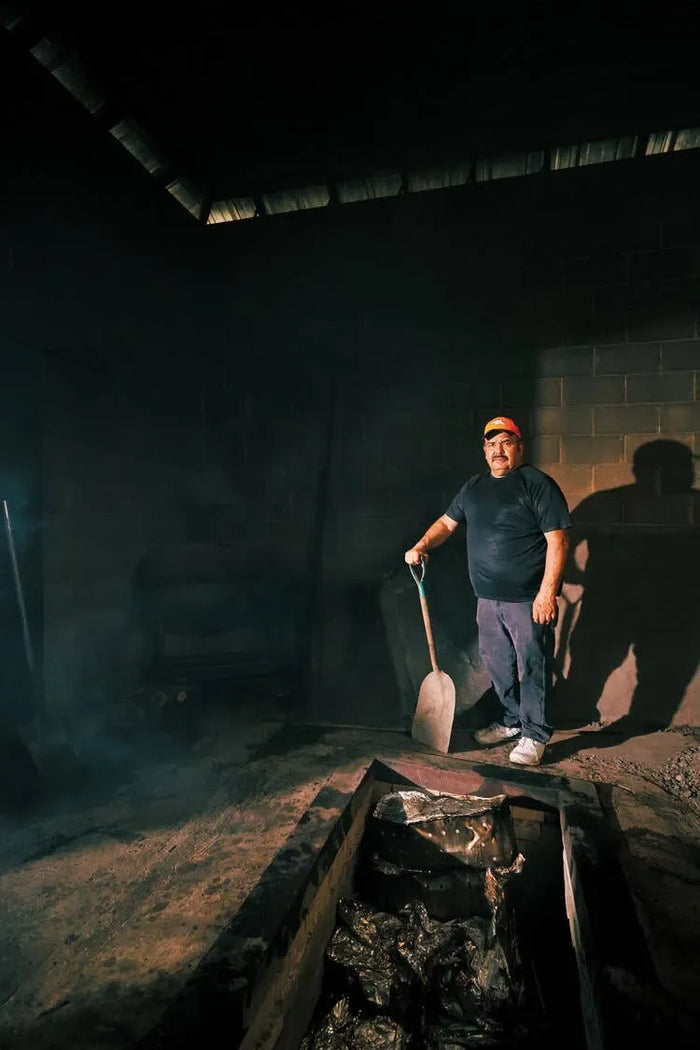 Man wearing a dark shirt and cap stands in a smokehouse beside a shovel and fire pit