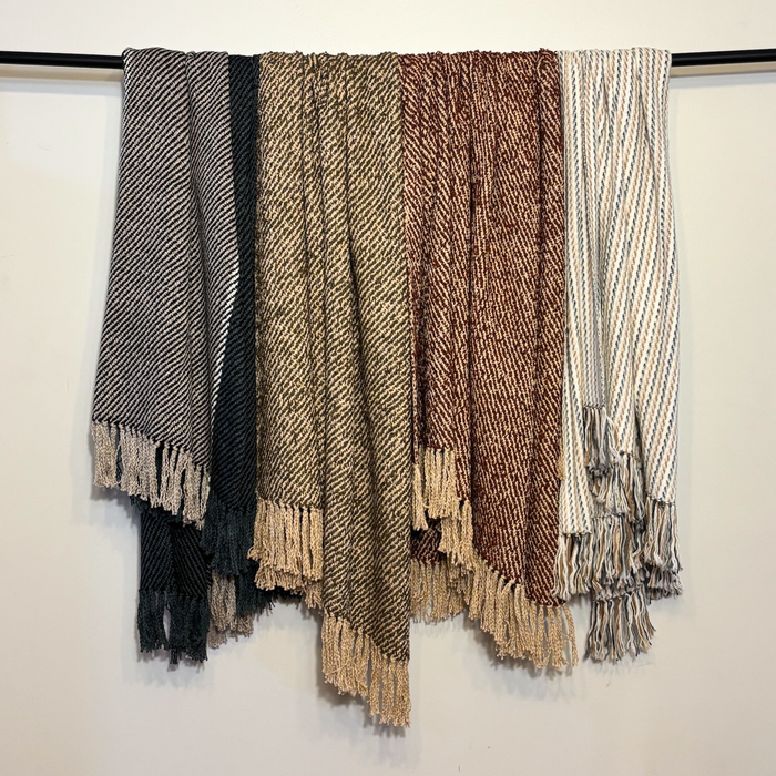Four woven throw blankets with fringe hanging from the bottom, draped side-by-side on a black rod against a plain white wall. The blankets feature different patterns and colors: black and gray herringbone, black and beige herringbone, brown and beige herringbone, and cream and gray stripes.