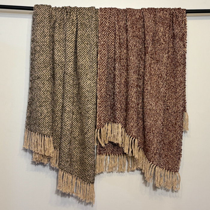 Two woven fringed throw blankets hanging from a black rod against a plain white wall. One blanket is heathered taupe and brown, and the other is a rich medium brown with beige fringes.