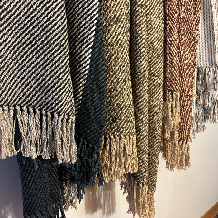 A close-up of several woven throw blankets hanging side by side, showing their textured herringbone patterns and fringed edges in neutral and earthy tones.