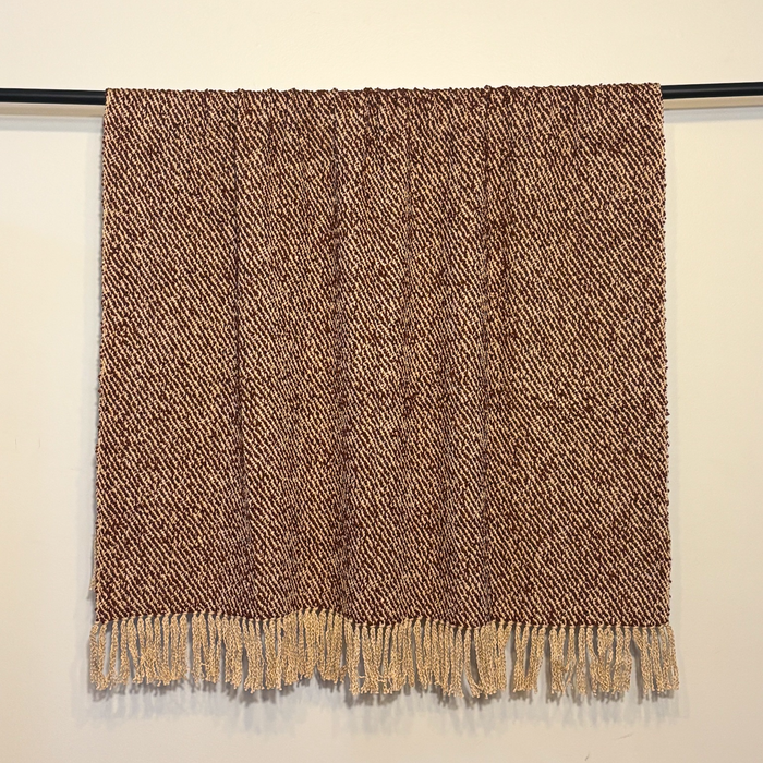 A brown and beige woven throw blanket with a textured herringbone pattern and fringed edges, hanging from a black rod against a plain off-white wall.