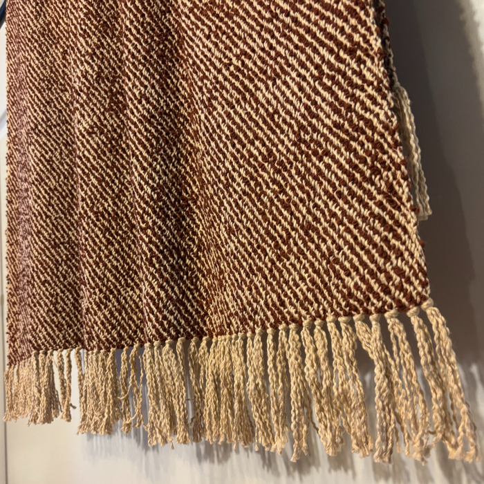 A close-up of a woven textile with a brown and beige herringbone pattern and fringed tassels along one edge, shown against a plain light background.