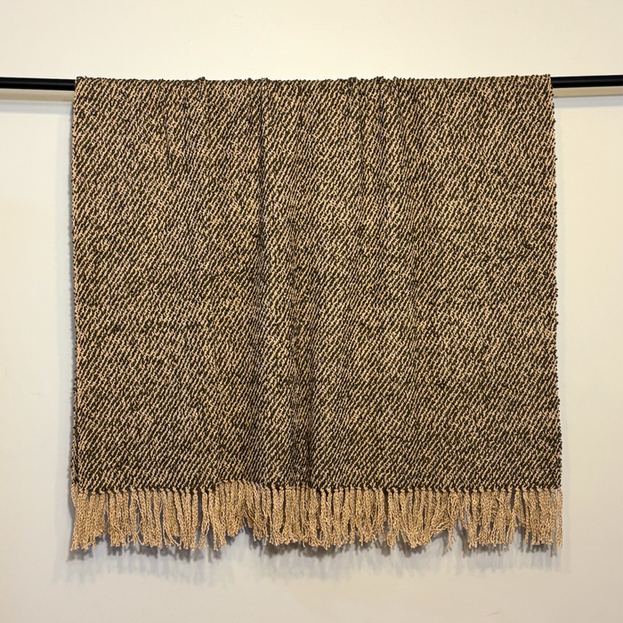 A woven throw blanket with a textured herringbone pattern in brown and beige, hanging from a black rod against a plain off-white wall.