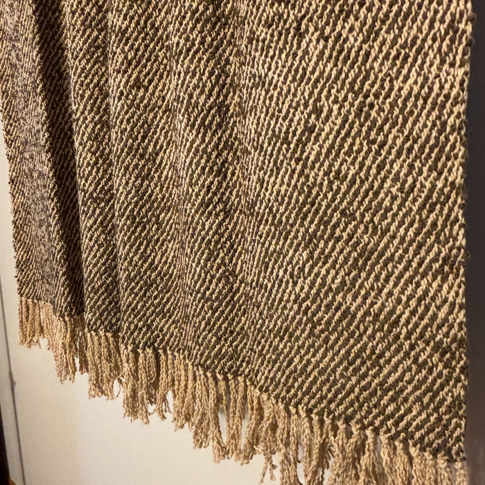 A close-up of a woven throw blanket with a textured herringbone pattern in brown and beige, featuring fringed edges.
