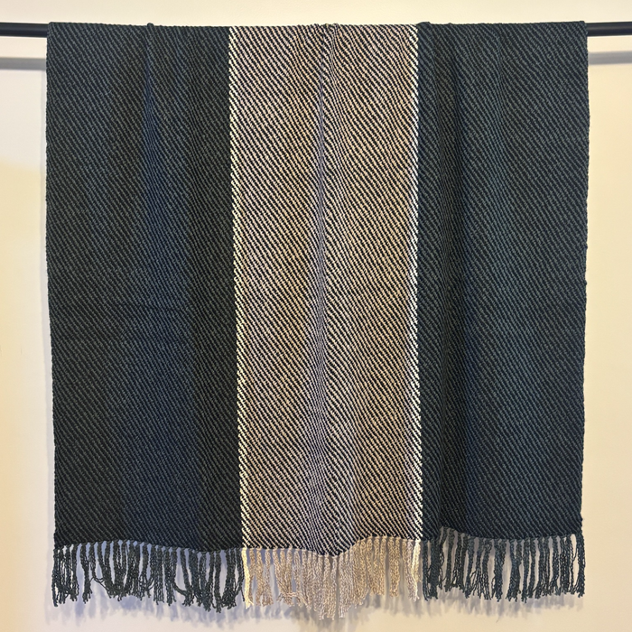 A woven throw blanket with fringe, featuring three vertical stripes in dark navy blue, a central beige and gray herringbone pattern, and dark navy blue sides, hanging from a black rod against a light beige wall.