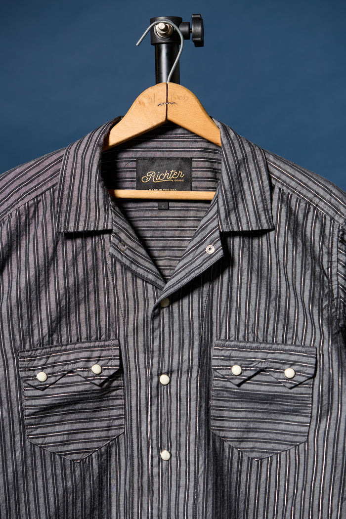 A gray and black vertically striped button-down shirt by Richter, hanging on a wooden hanger against a dark blue background.