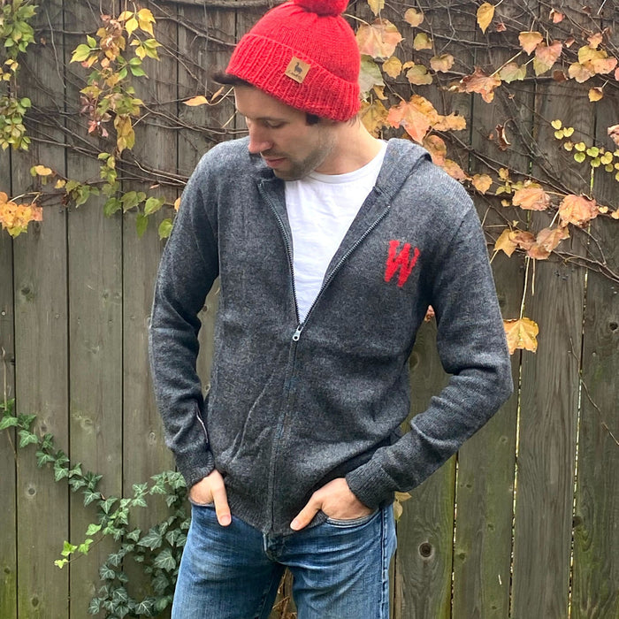Person wearing a dark gray hoodie with a red logo, red beanie, and blue jeans standing against a wooden fence with autumn leaves.